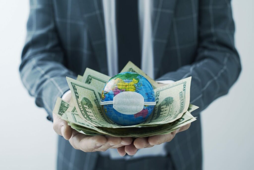 businessman holding a world globe with a protective mask on top of a handful of dollar bills