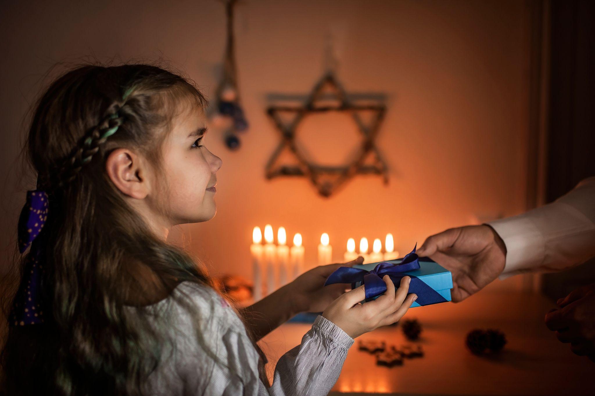Family celebrating Hanukkah, dad lighting candles on traditional menorah and giving gift to daughter