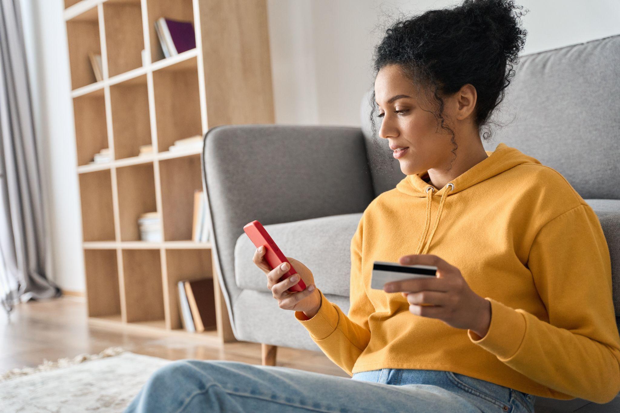 Young adult female consumer holding credit card and smartphone sitting on floor at home doing online banking transaction.