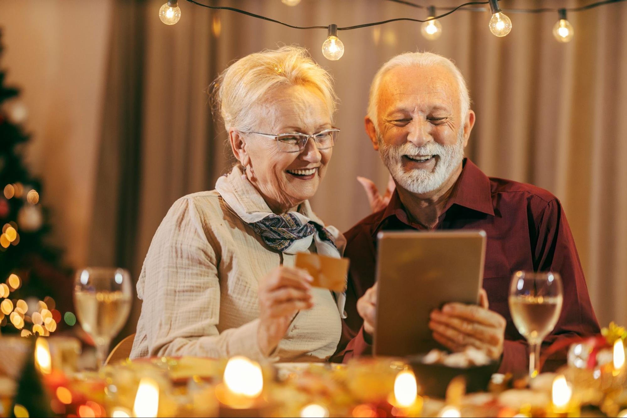 A couple holding a credit card and smiling while looking at a tablet. Holiday decorations are behind them.